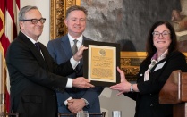 Zoom image: Three people standing indoors during an awards presentation. A framed award plaque is being presented to David E. Franasiak JD MBA &rsquo;78 by two presenters. All are dressed in formal attire, with an American flag and framed artwork in the background.