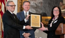 Zoom image: Three people standing indoors during an awards presentation. A framed award plaque is being presented to David E. Franasiak JD MBA &rsquo;78 by two presenters. All are dressed in formal attire, with an American flag and framed artwork in the background.