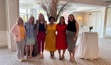 Six people stand side by side in a bright reception room, posing for a group photo in front of a tall decorative arrangement of branches with small lights. Cocktail tables and large windows are visible in the background. 