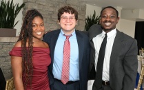 Three people standing together indoors at a formal event, dressed in elegant attire: one in a burgundy off-the-shoulder dress, one in a dark suit with a red patterned tie, and one in a gray suit with a black tie. Stone wall and plants visible in the background. 
