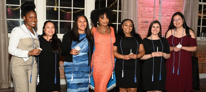 Eight students in formal attire stand together indoors, wearing blue and white honor cords and holding small items, posing for a group photo. 