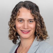 A person with short curly brown hair wearing a light gray blazer over a burgundy top, shown against a plain light gray background. 