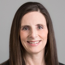 A person with long straight brown hair wearing a black blazer over a coral top, along with a simple necklace featuring a round pendant, posed against a plain gray background. 