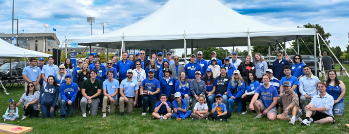 University at Buffalo community members gather for a group photo at an outdoor tailgate event under a large tent. 