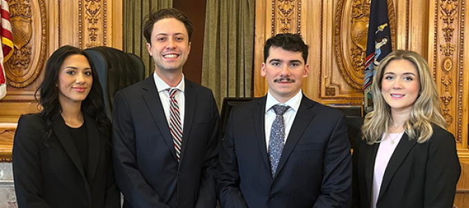 Four individuals in formal business attire standing in front of an ornate wooden judge’s bench inside a courtroom, with American and state flags on either side and the words ‘In God We Trust’ engraved above. 