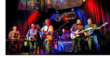 Live band performing on stage at Sportsmen’s Tavern, with colorful stage lighting and musical instruments including guitars, keyboard, and drums. 