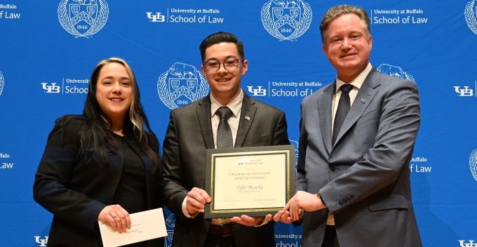 Three individuals stand in front of a University at Buffalo School of Law backdrop. The person in the center holds a framed award certificate, while the person on the left holds an envelope. The person on the right, dressed in a suit, stands beside them as part of an awards presentation. 