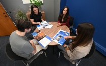 Four people sit around a small conference table in an office reviewing printed brochures and documents together, with additional papers spread across the table and plants and wall art in the background. 