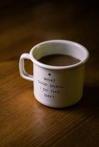 sitting on a wooden tabkle is a coffee mug that says 'what good shall i do this day??'. 