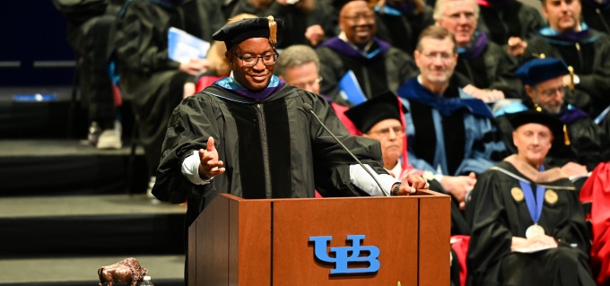 A speaker in academic regalia stands at a podium with the University at Buffalo logo during a commencement ceremony. Rows of faculty and graduates in caps and gowns sit behind the podium. 