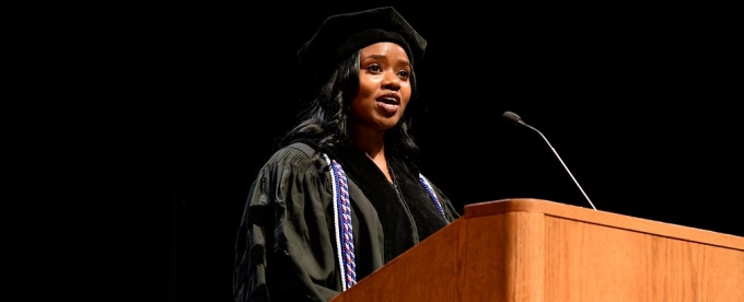 young woman wearing graduation regalia speaking at a UB podium. 