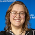 Sara Bachraty wearing glasses, black and white jacket, standing in front of a blue background. 